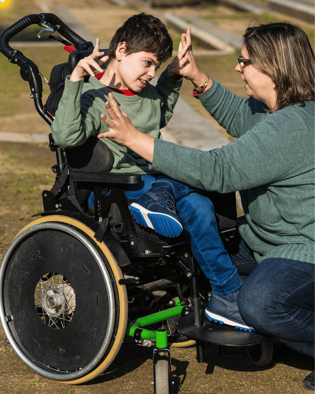 A child sitting in a wheelchair outdoors raises their hands while interacting with an adult kneeling beside them. Both are smiling and engaged with each other in a sunny park setting.