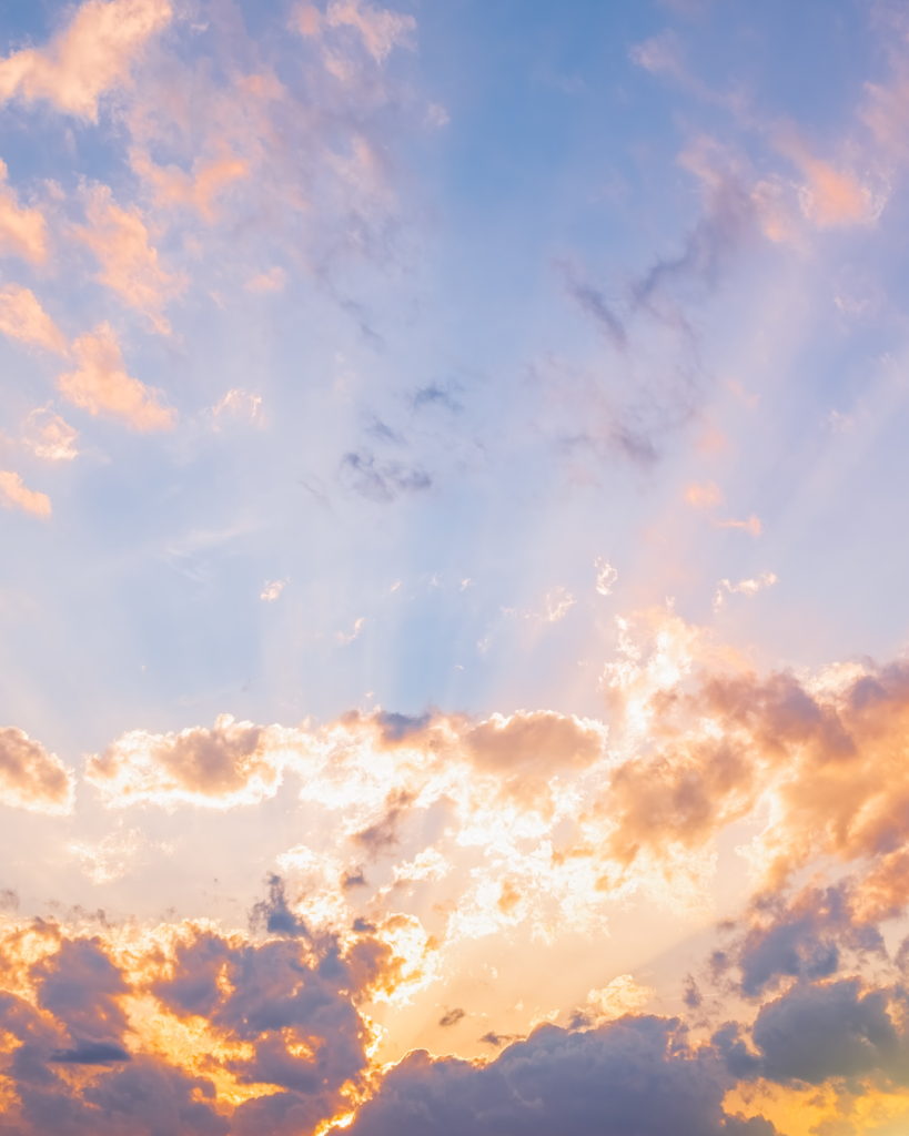 Sunset sky with golden sunbeams breaking through layered clouds in shades of orange, pink, and blue.