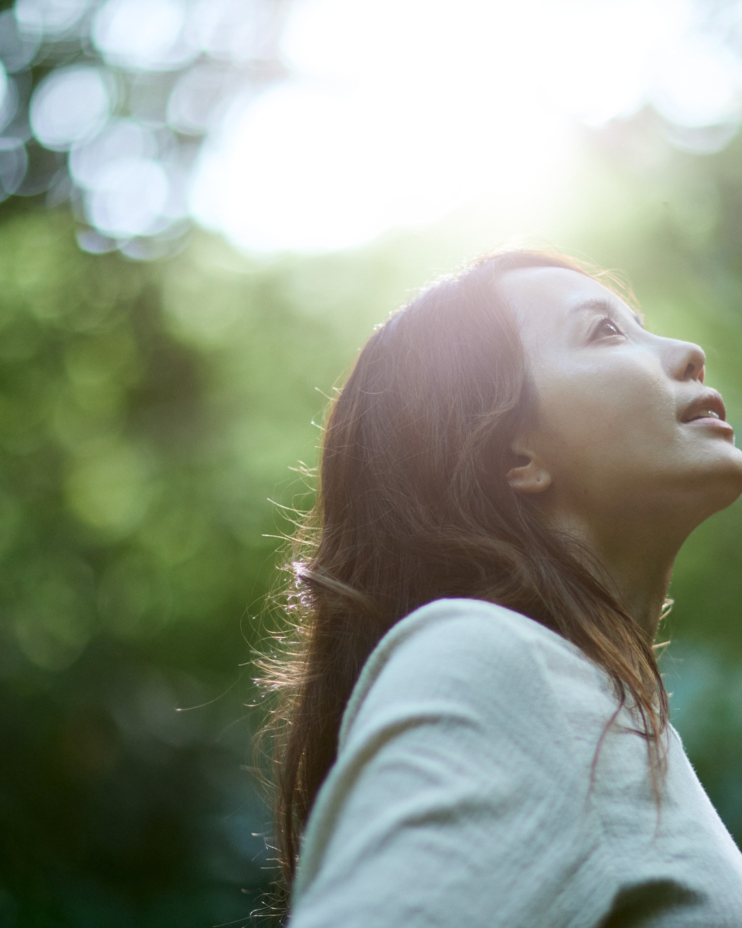 A woman with long brown hair tilts her head back to gaze upwards, her face lit by soft sunlight filtering through blurred green leaves.