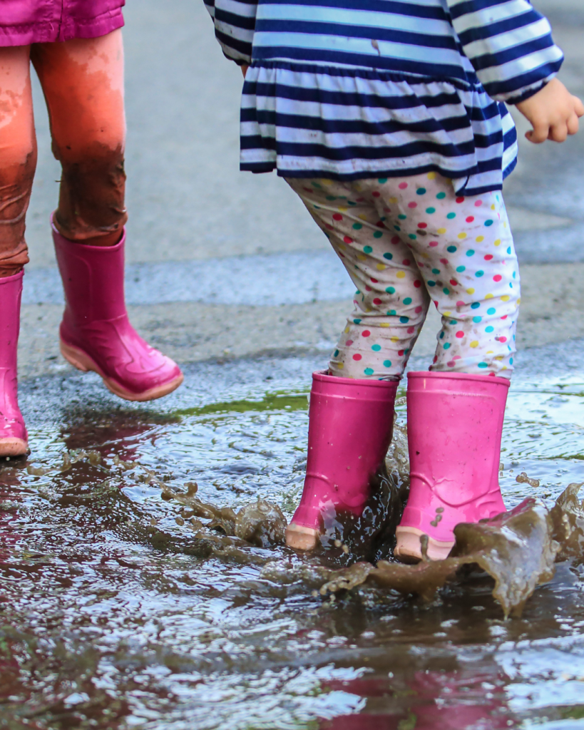 Two young children in pink wellies jump and splash in a muddy puddle, one wearing spotty leggings and a striped top, with water spraying around their boots.