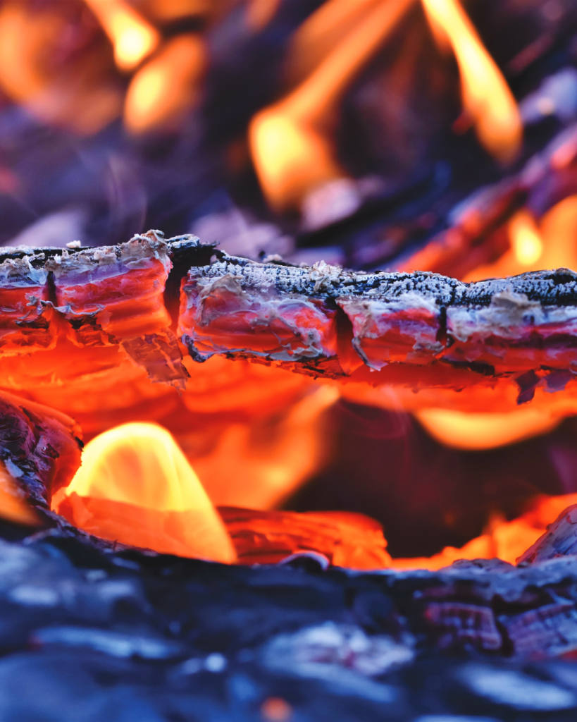 A close-up of glowing orange embers and small flames beneath a charred log, its edges crusted with pale ash.