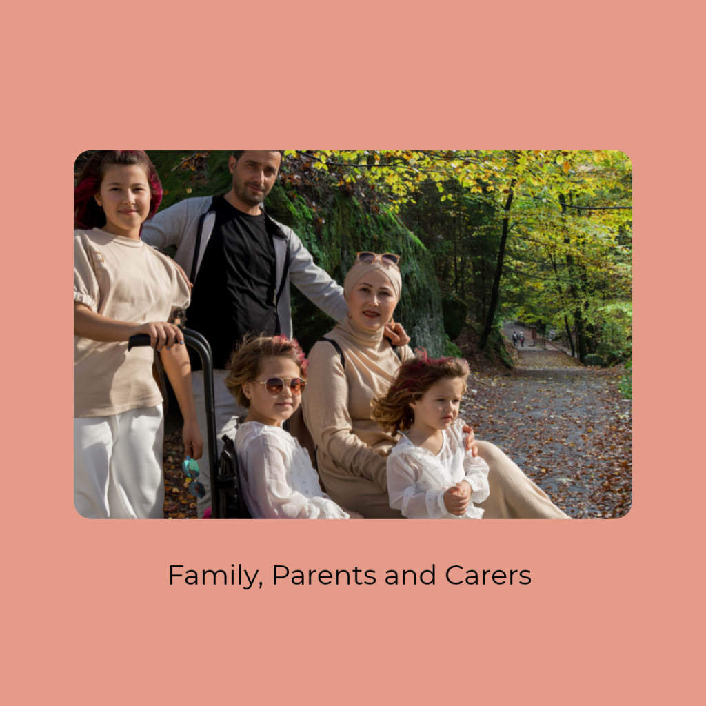 A family of five poses together on a forest trail surrounded by lush green and golden autumn foliage. An adult man and woman stand and sit with three young girls, one seated in a pink wheelchair. The family wears coordinated neutral and white outfits, with sunlight filtering through the trees and fallen leaves scattered along the path. 