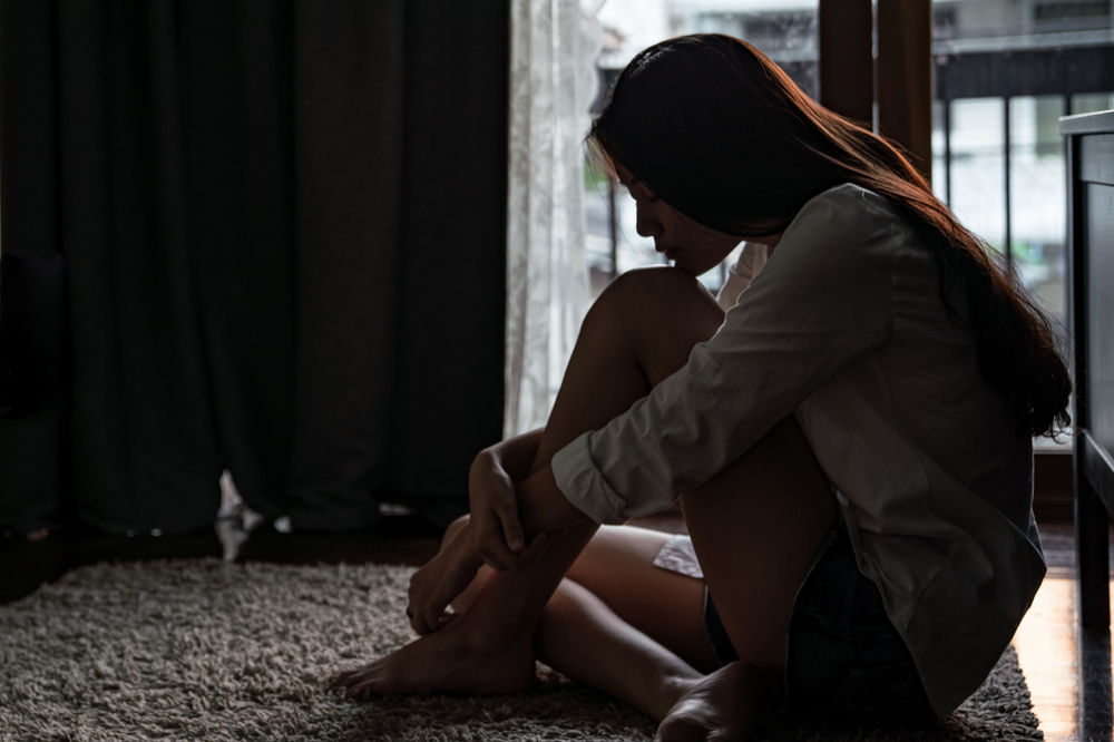 A young person with long hair sits on the floor by a curtained window, hugging their knees with their head bowed.