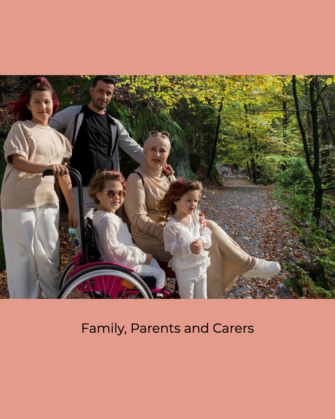 A family on an autumn woodland path under green and yellow leaves. An older child with red-streaked hair holds the handles of a bright pink wheelchair, where a younger child in sunglasses sits. One adult stands behind them, another adult in a beige headscarf sits beside them, and a toddler in white stands at the front. Caption: Family, Parents and Carers.

WHERE IT GOES (replacing the wooden FAMILY letters, media item 12413):
- autisticrealms.com/about/  (the For Families card)
- autisticrealms.com/family/
Nothing on morerealms.com uses the old picture.