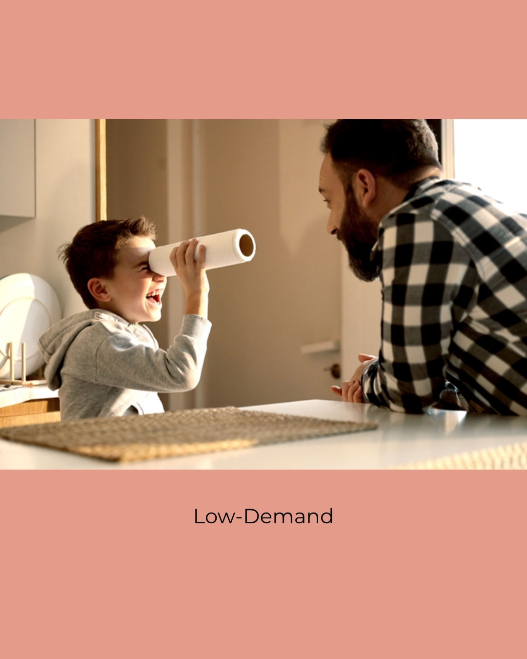 A laughing child looks through a paper towel roll at a smiling adult across a kitchen table. Caption: Low-Demand.