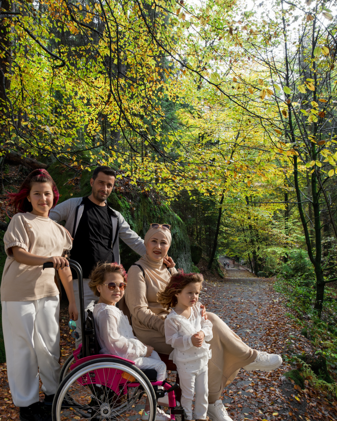 A family of five poses together on a forest trail surrounded by lush green and golden autumn foliage. An adult man and woman stand and sit with three young girls, one seated in a pink wheelchair. The family wears coordinated neutral and white outfits, with sunlight filtering through the trees and fallen leaves scattered along the path.